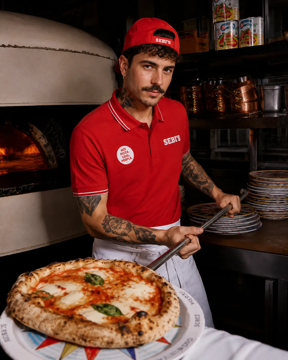 Pizzabäcker im roten Sebi&rsquo;s-Poloshirt zieht eine Margherita auf dem Schieber aus dem Holzofen